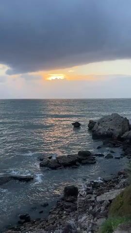 The video shows a rocky coastline and the ocean at dusk. The sky is cloudy with the sun setting behind the clouds, casting a warm glow. The waves are gently rolling onto the shore, and there are several large rocks scattered in the water. The scene is peaceful and serene.