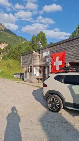 The video shows a scenic mountain landscape in Blenio, Switzerland. The camera is mounted on a vehicle, likely a car, as indicated by the view from inside and the presence of a Swiss flag on the side of a building. The setting appears to be a parking area or a clearing near a mountain hut or building constructed of wood.

The scene is bathed in bright sunlight, suggesting it is daytime, likely late morning or afternoon. The sky is blue with scattered white clouds. Lush green vegetation, including trees and grassy slopes, covers the mountainside. A winding dirt path is visible on the mountain.

Several vehicles are parked in the gravel area: a light-colored SUV with a Swiss flag attached, a white camper van, a blue car, a yellow car, and a red car. There are also some wooden posts marking parking spots. The overall atmosphere is peaceful and evokes a sense of outdoor adventure and relaxation.

Text visible includes signage on the building with arrows and what appears to be a hotel or restaurant name. The Swiss flag is a prominent detail.

The video is filmed vertically, which may suggest it was captured using a smartphone. The lack of visible people actively engaged in activities suggests this is a static scene, possibly a stop during a road trip or a visit to a mountain destination.