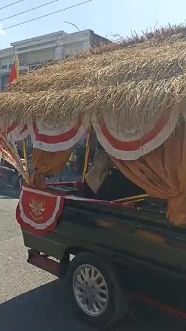 The video shows a street scene with several people sitting on the side of the road, some of whom appear to be watching something. There are also several motorcycles parked along the sidewalk. A building with signs is visible in the background. The scene appears to be a public gathering of some sort, possibly a parade or event. There is a flag pole with an Indonesian flag.