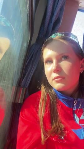 The video features a close-up shot of a young woman inside what appears to be a bus or train. She has fair skin, blue eyes, and light brown hair, and is wearing a red and blue jacket and a headband with decorative elements. The woman is looking towards the window, her expression somewhat serious or contemplative. Behind her is a blue curtain. The window shows a reflection of her face and suggests bright daylight outside. The overall setting suggests travel, and the lighting is bright and direct.