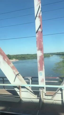 The video shows a view from a bridge overlooking a wide river under a clear blue sky. The bridge structure, made of metal beams painted white and red, is in the foreground, partially obscuring the view. On the far bank of the river, there is a dense green forest. Closer to the viewer, on the left bank, there appears to be a construction site with piles of sand or dirt and a large crane. The water is calm, with some patches of vegetation growing near the banks. There are no people visible in the frame. The overall impression is a sunny day with industrial and natural elements present.
