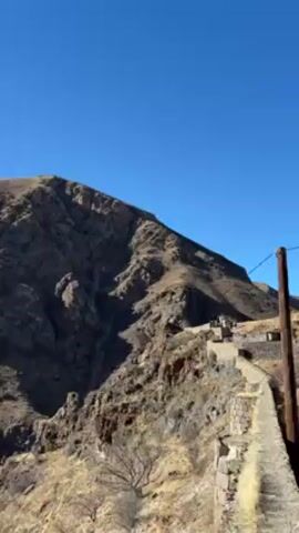 The video shows a rugged, rocky mountain landscape under a clear blue sky.  The mountain is mostly brown and gray, with visible layers and crevices. A pathway, likely made of stone, runs along the side of the mountain, with some stone structures visible. A few bare trees are visible near the bottom of the frame. The overall impression is one of a dry, mountainous region, possibly with historical significance due to the presence of the stone pathway and structures.