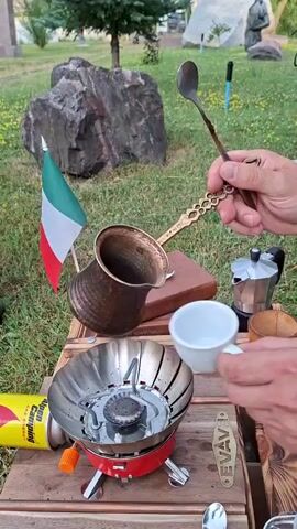 In an outdoor setting with green grass and large rocks, a person is pouring coffee from a copper cezve into a white cup. The cezve, with an ornate gold handle, is held over the cup, and dark liquid is seen flowing from it. The cup is positioned above a red portable camping stove. A can labeled "Camping" is visible on the left. A small Italian flag is planted in the ground behind the stove. To the right of the cup, a silver moka pot and a wooden object are present. The person's hands are holding the cezve and the cup. Another hand, holding a spoon, is visible higher up in the frame. The background includes trees and a statue.