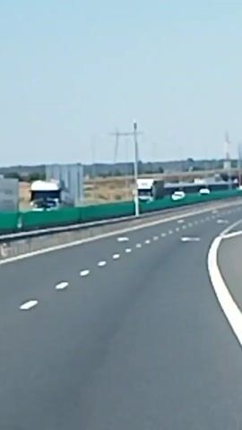 The video shows a highway with multiple lanes. The road is paved with asphalt and marked with white lines. There are lane markings and directional arrows on the road. On the left side of the highway, there is a green barrier and several vehicles, including trucks. In the distance, there are power lines and what appear to be industrial structures. On the right side of the highway, there is a metal guardrail and a green road sign with multiple directional arrows. The sky is clear and blue. The video appears to be filmed from a moving vehicle.