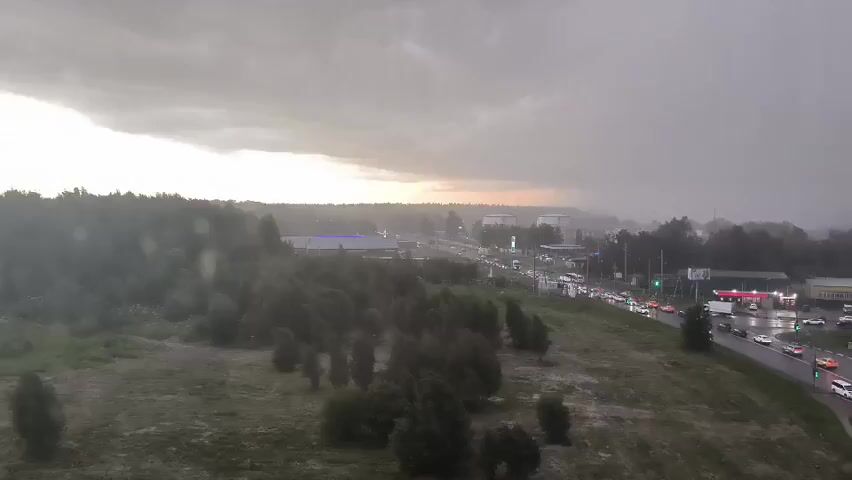 A wide aerial view shows a suburban landscape under a dark, stormy sky. In the foreground, a grassy hill slopes down toward a road lined with trees and buildings. The road is wet and reflects the lights of numerous cars, suggesting recent rainfall. The sky is overcast with heavy clouds, and a faint glow of sunset is visible on the horizon. The overall atmosphere is one of an approaching or ongoing storm.