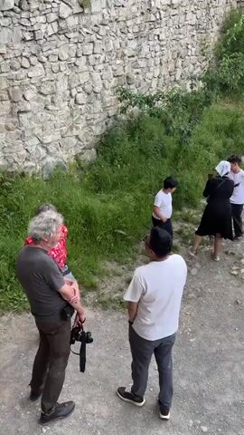 A group of people are gathered in an outdoor historical setting. The environment consists of ancient stone walls, greenery, and a dirt path. Some individuals are standing and observing, while others appear to be taking photos or interacting. There are children and adults present, dressed in casual clothing. The camera is positioned at a high angle, looking down at the scene.