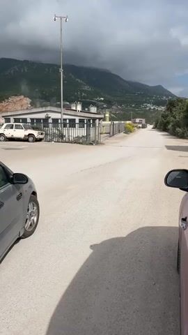 The video shows a silver Ford Focus station wagon driving away from the camera down a dusty, white dirt road. It accelerates, emitting a large cloud of black smoke from its exhaust. The car drives past a white industrial building with a metal fence surrounding it. In the distance, there is a view of mountains under a cloudy sky. The camera is positioned inside a red car, with part of the car’s side mirror visible in the right side of the frame.