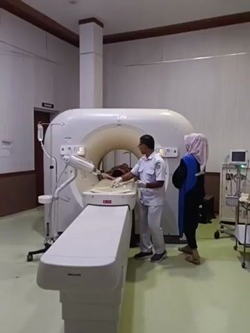 The video shows a medical imaging room. A patient lies on a bed that is part of a CT scanner. A medical professional in scrubs stands next to the patient, appearing to adjust something or check on them. Another person, possibly a technician or a second medical professional, stands to the right of the scanner, observing. The CT scanner is a large, white, circular machine. The room has a pale green floor and light-colored walls. Medical equipment, including a monitor on a cart, is visible in the background. The lighting is bright and clinical.