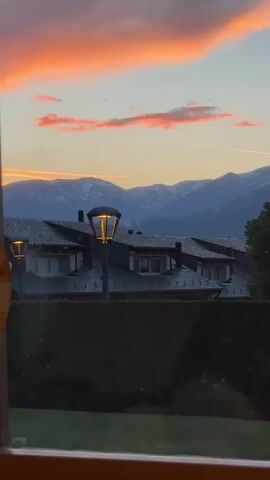 The video shows a scenic view of mountains at sunset from a window. In the foreground, there's the dark roof of a building with dormer windows and a few chimneys. Two vintage-style lampposts stand illuminated in front of the roof.  A green hedge runs along the bottom of the frame, obscuring the immediate foreground. Beyond the roof and lampposts, a range of mountains rises in the distance, partially covered in snow. The sky is filled with pink and orange hues from the sunset, with streaks of white clouds near the horizon. The video seems to have been taken from indoors, as a window frame is visible at the bottom. There is audible speech in the background.