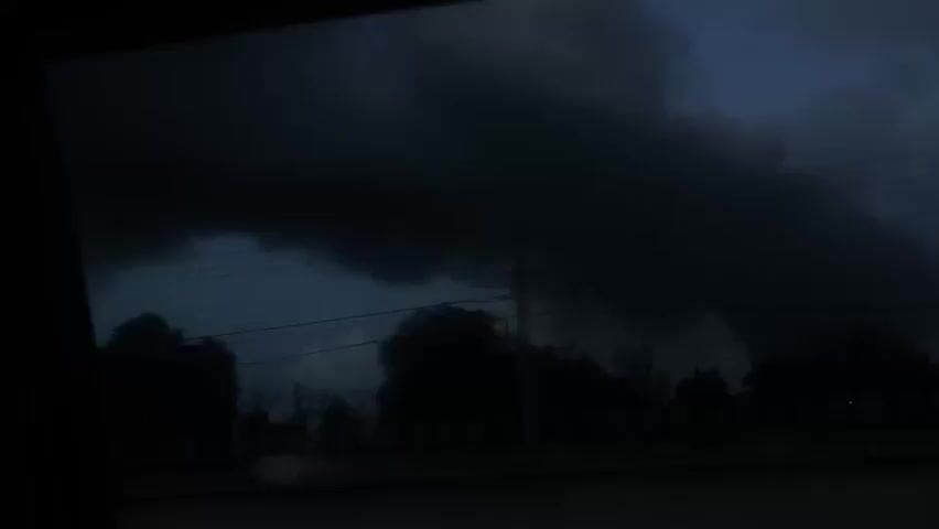 The video, shot from a moving vehicle, shows a dark and ominous sky with heavy, low-hanging clouds. The foreground features silhouettes of trees, including palm trees, and glimpses of buildings illuminated by streetlights. A white pickup truck passes by on the road. The overall atmosphere is dark and foreboding, suggesting an approaching storm.