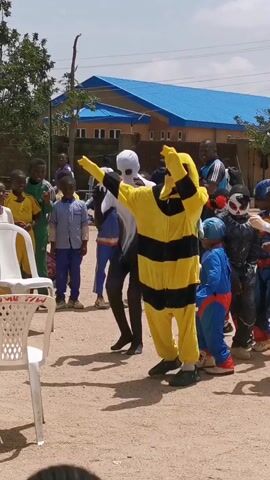 Here is a concise description of the video:

The video captures an outdoor gathering, likely a children's event, featuring several people in costume. A person in a yellow and black bee costume and another in a white alien costume are dancing together, surrounded by children dressed as superheroes. The location appears to be a schoolyard or similar open area, with a blue-roofed building in the background. Several white plastic chairs are scattered in the scene.