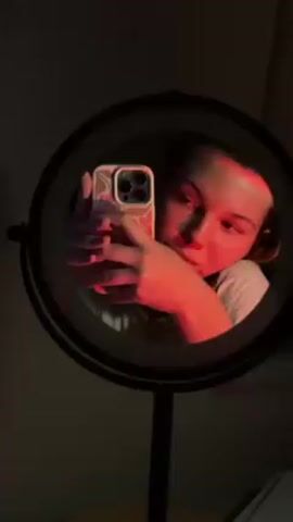 In the video, a young woman is recording herself using a smartphone. She holds the phone, which has a white case with a pink floral design, up to a round mirror. The light source illuminates her face in red, creating striking contrasts. The camera captures a close-up of her face, focusing on her eyes.