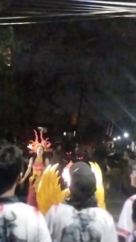 The video shows a nighttime outdoor event, likely a parade or festival. In the center, a person in a vibrant costume resembling a sun or fire goddess is walking. The costume features a headdress with flame-like elements, and a shimmering outfit. Behind and around this central figure, other participants are visible, some also in costume with large, flame-shaped props. A crowd of onlookers is present, facing the parade. Some individuals in the foreground are wearing white t-shirts with a graphic of a bull. The environment appears to be a street with trees and overhead wires, suggesting an urban or suburban setting. The lighting is dim, with some artificial lights from the event and streetlights visible. The overall atmosphere seems festive and celebratory.