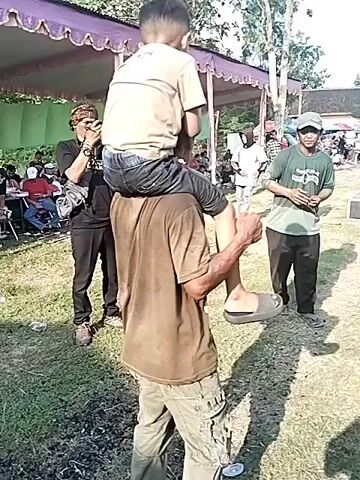 The video shows a man carrying a young boy on his shoulders. They are standing on a grassy field, in front of a large tent structure. The man is wearing a brown t-shirt, camo pants, and brown sandals. The boy is wearing a tan t-shirt and blue jeans. There are people standing around them, some are seated at tables under the tent. A man wearing a black shirt and a bandana is taking pictures. The camera turns to show a group of people dancing to music in the background. The setting is outdoors and appears to be some kind of event or gathering.