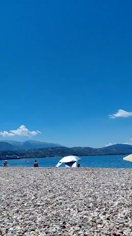 The video captures a wide shot of a pebble beach on a clear, sunny day. The sky is a vibrant blue with a few wispy clouds in the distance. The sea stretches out towards a range of hazy mountains on the horizon. In the foreground, people are scattered across the beach. Some are sitting or standing, while others appear to be in or near the water. There are also beach umbrellas, one of which is white and dome-shaped, and another made of straw. The overall atmosphere is one of a relaxed day at the beach.