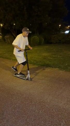 A person wearing a white t-shirt, white shorts, black knee pads, a grey baseball cap, and black and white sneakers is riding a silver and black scooter on a paved path at night. The person is in a park with grass and trees in the background. The person is moving from left to right in the frame, bending their knees and leaning back as they ride. The lighting suggests streetlights or distant building lights are illuminating the scene.