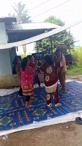 A group of five people, four women and one man, are dancing on a large blue and green patterned mat. The women are wearing colorful traditional clothing. The man is wearing a light brown shirt and khaki pants. They are under a white canopy, which is supported by a light blue pole. Behind them, there is a gray metal roof and a green bush. There are two large speaker boxes on the right side of the frame. The ground is dirt and there are some plastic bottles on the edge of the mat. The overall scene appears to be an outdoor gathering.