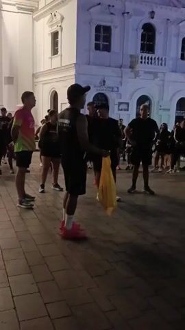 A group of approximately 20 people are gathered on a cobblestone plaza in front of a large, white building illuminated with purple light. The people are mostly dressed in dark athletic clothing, some with brightly colored running shoes. One person in the foreground is holding a yellow bag. They appear to be listening to someone who is standing near a black speaker. The building in the background features arched windows and a prominent facade. The overall setting suggests an organized group activity, possibly a running club or tour, taking place in an urban environment at night.