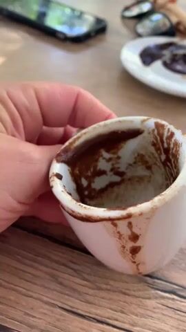 A close-up shot shows a person's hand holding a white coffee cup. The inside of the cup is coated with dark brown coffee grounds, forming patterns. The cup is held over a wooden surface, possibly a table. In the background, out of focus, is a white saucer with more coffee grounds, a mobile phone, and a pair of glasses. The lighting suggests an outdoor or well-lit indoor setting.