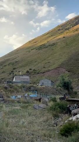 The video shows a mountainous landscape with a gentle slope covered in grass and some rocky patches. In the mid-ground, there are two small buildings. The building on the right is a simple, grey, rectangular structure with a window and a door. In front of this building, two people are visible. Both are wearing white protective suits, suggesting they might be beekeepers. They appear to be engaged in an activity near some colorful boxes, which could be beehives. The person on the left is standing and facing the person on the right, who is bent over the boxes. The person on the left is holding a tool or object. The overall impression is of a rural, possibly agricultural setting. The sky above is partly cloudy, with the sun illuminating the side of the mountain. The foreground consists of dry grass and rocks.