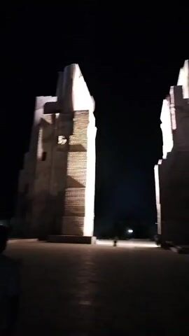 This media file captures a nighttime scene in Shahrisabz City, Uzbekistan, featuring the impressive remains of ancient structures. The primary subjects are two towering, partially ruined brick edifices, likely remnants of historical significance, illuminated by spotlights that cast dramatic shadows. The structures have a stepped, architectural design, with visible brickwork creating horizontal lines.

The scene is set in an open, paved plaza. In the foreground, the back of a person wearing a dark t-shirt is visible, suggesting someone is observing or walking through the area. Further back, between the two illuminated structures, a few other individuals are present, appearing as small silhouettes against the light, possibly tourists or locals enjoying the illuminated site. One person is walking towards the camera, while a small group is gathered near the base of one of the structures.

The time of day is clearly night, indicated by the dark sky and the artificial lighting on the monuments. There is no visible text in the media file. The overall mood is one of historical grandeur and nighttime illumination, highlighting the ancient architecture against the dark expanse of the night. The emptiness of the plaza and the solitary figures contribute to a sense of awe and perhaps a touch of mystery.