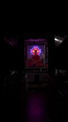 The video shows a dark interior with a statue of Buddha in the center, illuminated by a colorful, circular light display behind it. The light display cycles through various colors, including pink, purple, green, and yellow, creating a halo effect around the Buddha. The Buddha statue is seated in a meditative pose and appears to be made of a reddish material. There are floral arrangements on either side of the statue, and a small offering is placed at its base. The surrounding area is dark, with the only light emanating from the display. The overall impression is one of tranquility and reverence.