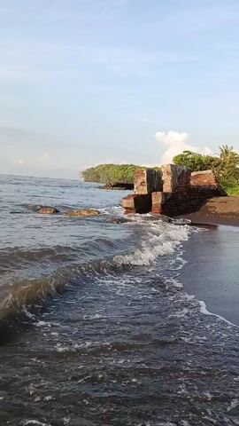 Here is a concise description of the video: 

The video captures a view of a beach on a slightly cloudy day. The sand is dark, and gentle waves are lapping at the shore. In the distance, there's a small, tree-covered headland. In the foreground, there are remnants of brick structures partly submerged in the water, suggesting ruins or old construction along the coastline. The focus is on the interplay of the waves and the static brick remains, creating a scene of natural erosion and history.
