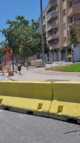 The video shows a street-level view of an urban environment, likely in a city. Initially, the camera is positioned behind a row of red and white plastic traffic barriers lining a road. A pedestrian walks into the background.  The camera then moves, apparently onboard a vehicle traveling along tram tracks. The shot reveals an empty tram stop with overhead power lines. As the vehicle progresses, the camera pans up to show a pair of tall, modern-style buildings, and then a construction crane. The sky is clear and bright throughout the video, suggesting a sunny day. The overall impression is a blend of urban infrastructure with ongoing construction and public transportation facilities.