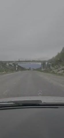 This is a first-person view from inside a car driving down a paved road in Kyrgyzstan. The road is in moderate condition, with visible wear and tear. On either side of the road, there are grassy, rocky slopes with sparse vegetation and trees. In the distance, mountains are visible under a cloudy sky. An overpass stretches across the road, supported by concrete pillars. A white vehicle is seen in the distance on the road, ahead of the car from which the video is filmed. The sky is overcast, suggesting a cloudy or possibly rainy day. The overall mood is one of travel through a somewhat rugged, natural landscape. There is a Mercedes-Benz emblem visible on the hood of the car in the foreground. No people are clearly visible, but the activity is driving.