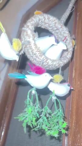 A close-up shot shows a wind chime hanging from a wooden door. The wind chime is made of a woven rope circle with small, white bird figures attached. Some of the birds have colorful feathers on their heads and tails. Small, green artificial plants hang from the bottom of the birds. The wind chime is swaying gently, suggesting a slight breeze. The lighting is dim, and the background is blurred, focusing attention on the wind chime.