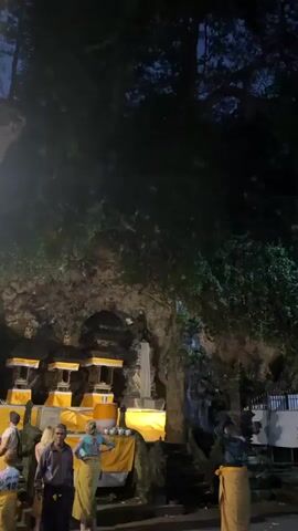 The video shows a nighttime scene at a temple complex. A large, dark tree with many leaves dominates the upper portion of the frame, with the night sky visible through its branches. Below the tree, a dark cave or rock face is visible on the left, with illuminated shrines decorated with yellow fabric. Several people are present. On the left, three individuals are standing. One appears to be a man in a blue shirt and a sarong, talking to a woman in a white top and a blonde woman in a light-colored top and a sarong. To the right of the cave, a multi-tiered pagoda-style temple structure is visible, adorned with gold trim. Another person, dressed in a sarong, stands to the right of this structure, facing away from the camera and seemingly observing the scene. The overall atmosphere is dark, with artificial lighting illuminating the temple structures and the immediate vicinity. Some faint, blurred objects resembling flying animals, possibly bats, are visible in the air. The scene appears to be a cultural or religious gathering.