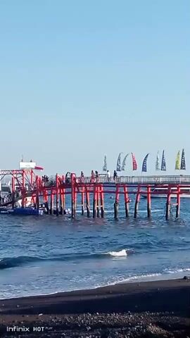 The video shows a view of a pier with red torii gates extending over the water, leading to a larger structure at the end. People are visible walking on the pier and on the structure. Several flags are flying from poles on the pier. The water is blue and appears to be the sea, with gentle waves lapping at a dark-colored beach in the foreground. The sky is clear and blue. There is a watermark in the bottom left corner that reads "Infinix HOTPlay".