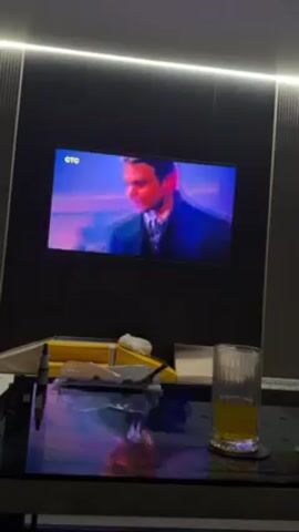 A video shows a TV on the wall playing a scene from a movie, possibly "The Thomas Crown Affair." The TV screen displays a conversation between two men at a bar or restaurant, one of whom may be Pierce Brosnan. In the foreground, there's a black, reflective coffee table. On it are a glass of orange juice or similar drink, some papers and objects, and a yellow item that is possibly a car. The room has a modern design with a dark wall behind the TV and LED lighting strips on the ceiling. The overall lighting is dim and the reflection on the coffee table creates a blue hue.