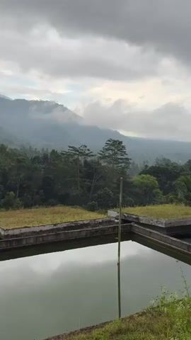 The video shows a cloudy sky over a lush green mountainous landscape. In the foreground, there is a large rectangular reservoir filled with murky green water. The reservoir has concrete walls and is partially covered by rectangular concrete platforms with grass growing on them. A thin vertical pole, possibly a flagpole, is visible in the water on the right side of the reservoir. The mountains in the background are covered in dense forest and are partially obscured by mist. The overall impression is one of a natural, somewhat undeveloped, and tranquil environment.