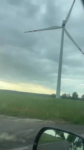 The video shows a wind turbine in a field with overcast skies. The turbine is white with red accents on the tips of its blades. The field is green with tall grass and there are trees in the distance. A grey paved path with a green and white barrier is visible in front of the turbine. The video is taken from a moving vehicle, with the side mirror visible in the bottom left corner.