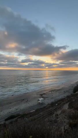 A static, high-angle video captures a coastal scene at dusk. The ocean dominates the left side of the frame, with gentle waves lapping at a sandy beach. Wispy clouds streak across the sky, tinged with orange and yellow from the setting sun, which is a bright spot on the horizon. A lifeguard tower stands on the beach. In the foreground, a wooden railing and dry grass create a natural border, with three small figures walking down a path on the left.