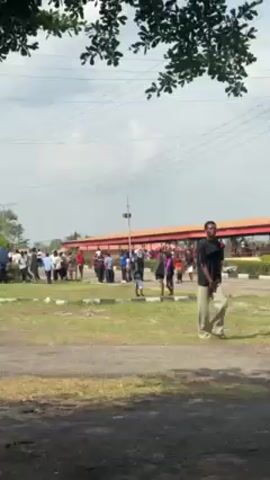 The video shows a group of people standing outdoors in a grassy area near a building with a red roof. Some of the people are carrying umbrellas. There are utility poles and overhead wires in the background. A few people are walking in the foreground. The sky is cloudy.