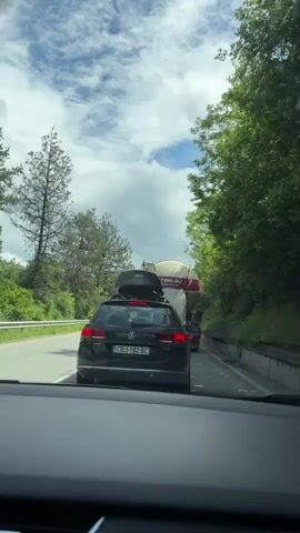 The image captures a first-person perspective from inside a vehicle, looking ahead at traffic on a road. Directly in front, a black Volkswagen Passat station wagon is visible, equipped with a black Thule roof box. The Passat's rear features a Bulgarian license plate displaying "CB 5182 BC," along with "PASSAT" and "2.0 TDI" badging. Its brake lights are illuminated, suggesting the vehicle is either stopped or moving slowly in a queue.

Behind the Passat, a large white tanker truck, likely a cement or liquid transport vehicle, can be seen. Prominently displayed on its side are the words "TSELEV" in red, "VOLVO," and the phone number "0888 372 373."

The road is bordered by dense green foliage and tall trees on both sides, with a metal guardrail visible on the left. The sky overhead is partly cloudy with patches of blue, indicating a bright daytime setting. The overall scene depicts a traffic situation on a road winding through a verdant landscape in Bulgaria, consistent with the provided location context of Kostandovo. The vehicles appear to be in a temporary halt or slow-moving traffic.