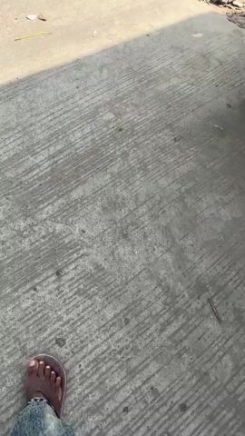 The video opens showing a concrete surface with some debris. The camera then pans to reveal a street scene with people, vehicles, and buildings. Several individuals are visible, some walking and others standing. There are cars and bicycles present, as well as shops lining the street. The environment appears to be an outdoor urban setting, possibly a market or a busy street. The lighting suggests it is daytime, with shadows indicating sunlight. The footage is shaky and the camera angle is sometimes tilted, giving a dynamic perspective of the scene.