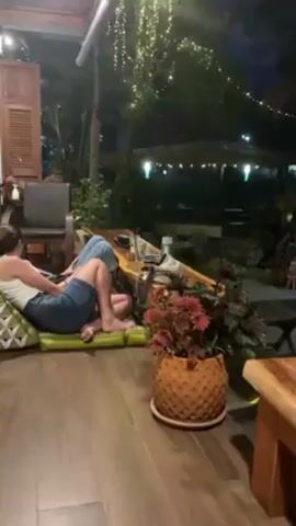 The video showcases an outdoor patio area at night, dimly lit by string lights and various lamps.  A person is lounging on a cushioned bench in the foreground. A wooden table and a potted plant with patterned, brown, pineapple-like designs are also in the foreground. The patio area features multiple seating areas with wooden furniture and cushions.  Some structures resemble small treehouses.  Vegetation is present throughout the scene.  In the background, there's a building with a roof and what appears to be a stained-glass window.  The overall atmosphere suggests a relaxed and casual setting.