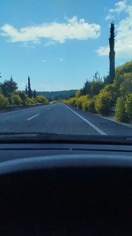 The video is filmed from the perspective of a passenger in a car, looking out the windshield. The car is driving on a highway. The road is asphalt and has white lane markings. On either side of the road are green trees and bushes, with some yellow flowers visible on the bushes on the right side of the road. The sky is blue with some white clouds. There is another white car visible in the distance on the highway. The interior of the car, specifically the dashboard, is visible in the foreground.