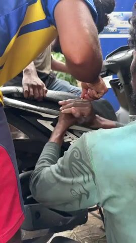 The video captures three men repairing a black Honda Dio scooter in an outdoor, daylight setting. In the foreground, a man wearing a blue and yellow striped t-shirt and blue and red shorts leans over the scooter from the left side, with his back mostly to the camera. To his right, another man, dressed in a teal long-sleeved shirt and dark pants, kneels beside the scooter. He has dark, curly hair tied back and is actively working on the area around the scooter's seat, using a small tool with both hands, applying significant force as if prying or manipulating a component. A third man is partially visible in the background, sitting on the scooter itself, observing the repair.

The scooter is black with white graphics and the "Dio" logo visible on its side panel. The ground beneath them is dirt or gravel. In the background, a blue corrugated metal sheet serves as a wall, and a white sign with blue text, which is illegible, is affixed to it. The overall activity appears to be a collaborative manual repair effort on the scooter's seat or the compartment beneath it. The men display concentration on their task. The scene suggests a casual repair workshop or a roadside service point during bright, clear weather.