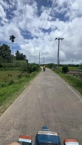 The video shows a first-person point of view from a blue Honda motorcycle driving on a narrow concrete road. Another person on a yellow motorcycle is ahead and receding into the distance. Lush green foliage lines the sides of the road, bordering fields of green on both sides. Power lines stretch overhead, supported by tall utility poles. The sky is partly cloudy with a bright sunny day.