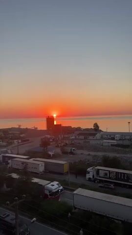 A time-lapse video captures a sunset over a body of water. In the foreground, there is a road with several semi-trucks traveling on it. In the middle ground, there are industrial buildings, construction equipment, and more trucks. The sun is low on the horizon, casting a warm orange glow across the sky and reflecting on the water. The sky transitions from a pale blue to a deep orange and pink hue as the sun sets.