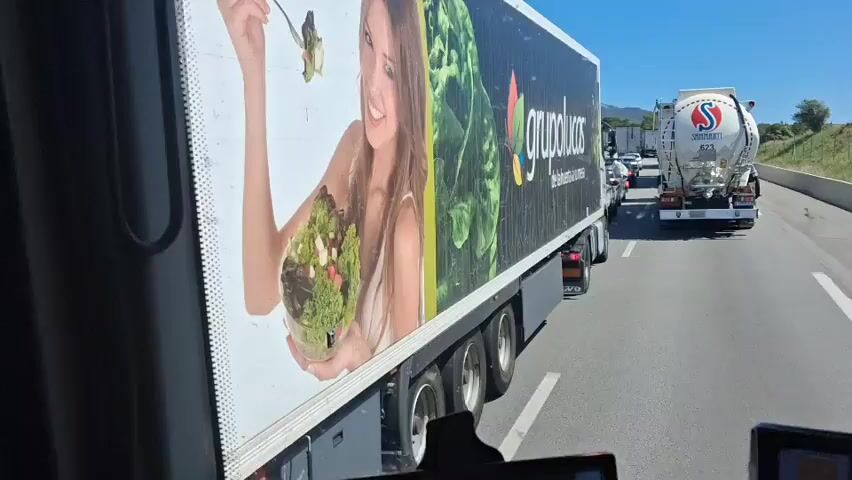The video, shot from inside a vehicle, shows a traffic jam on a highway on a sunny day. The vehicle appears to be a truck itself, sitting in heavy traffic. The truck in the next lane has a large advertisement for "grupolucas" on its side, featuring a woman holding fresh produce.  Several cars and trucks are visible in the lane ahead, many sitting stationary. A tank truck with the word "Shanahan" on it is directly in front of the filming vehicle. The windshield is partially visible in the frame.  A small screen is visible in the lower part of the video, perhaps showing GPS navigation. A radio is playing music in the background.
