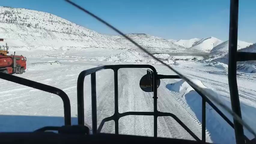 The video shows a vehicle's perspective driving on a snow-covered road within a mining landscape. To the left, a large, red tank truck is parked next to a yellow dump truck. The road is covered in tracks, and the surrounding environment features snow-covered hills and mountains under a clear blue sky. The view is framed by the safety bars of the vehicle's cab.