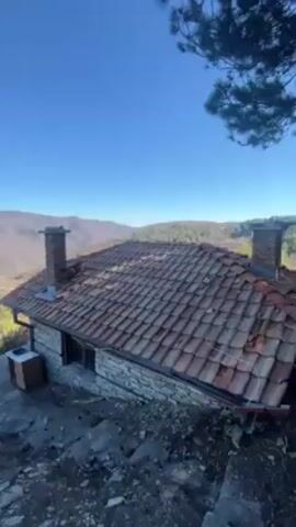 The video shows a small stone house with a red tile roof in a mountainous landscape. The house has a chimney and a small window. Steps lead down from the house. The landscape is a valley with trees and brown vegetation. The sky is blue and clear. A hand with painted nails appears in the frame momentarily. The overall impression is a remote and peaceful setting.