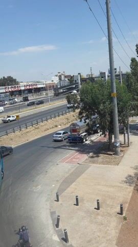 The video shows a view overlooking a multi-lane highway in an urban area on a sunny day. Cars and trucks are actively traveling in both directions. There is a pedestrian walkway visible alongside the road. On the side of the highway, there are various businesses and billboards. A bus is partially visible in the foreground. At the bottom of the video, a person on a motorcycle is seen. The sky is blue with scattered clouds.