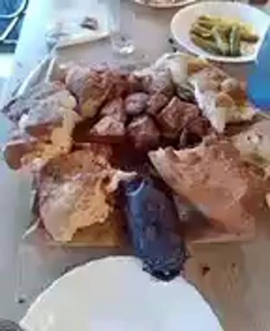 A group of people are gathered around a table, which is laden with food. There are plates of meat, bread, and what appears to be pickles. A fork is resting on a white plate in the foreground. One person is seen with their hand reaching for food on the table. The scene appears to be a casual meal.