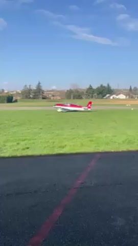 The video shows a small red and white plane sitting on a grass field adjacent to a cracked tarmac. The sky above is blue with scattered clouds. In the background, there are trees, houses, and fields. The plane appears stationary in the middle of the frame. The camera's perspective is from the edge of the tarmac looking across the grass towards the plane.